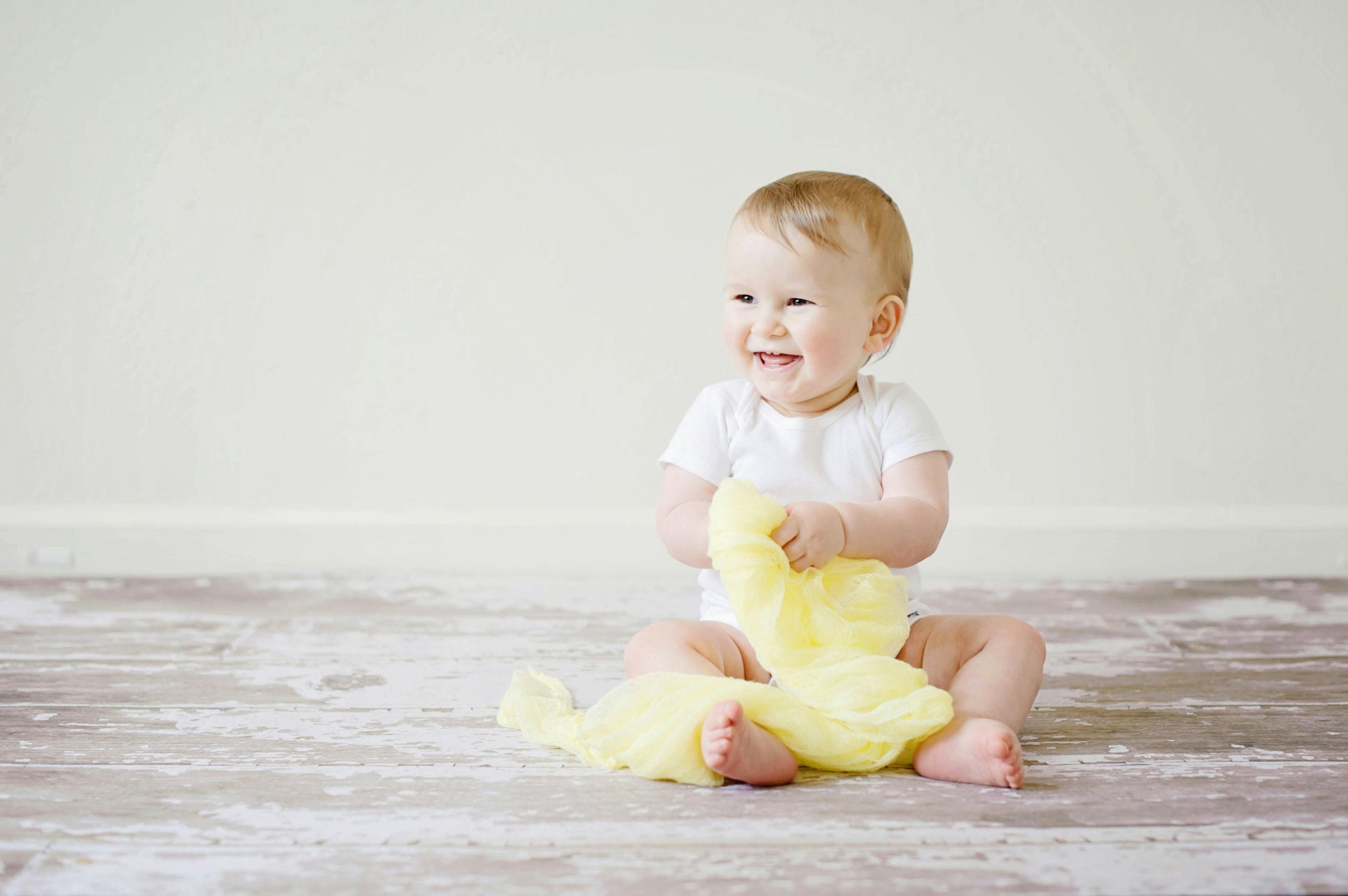 Baby sitting on a wooden floor holding a yellow toy against a white background